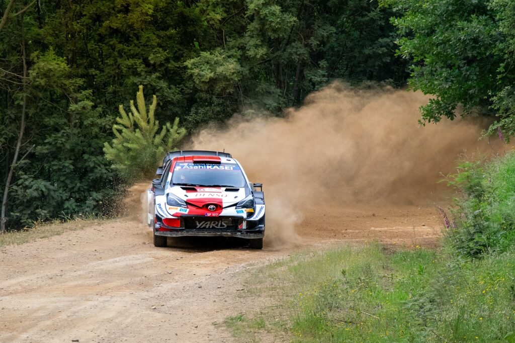 Kuvassa Kalle Rovanperän Toyota Yaris WRC -ralliauto ajaa hiekkatiellä metsän keskellä, nostattaen ilmaan suuria pölypilviä. Auto on punavalkoinen ja siinä on näkyvillä sponsoreiden logot, kuten DENSO ja Asahi KASEI. Taustalla näkyy tiheä metsä ja vihreää kasvillisuutta.